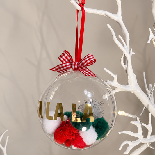 Clear ornament with red, green, white pom-poms and gold lettering, hanging from a white branch.