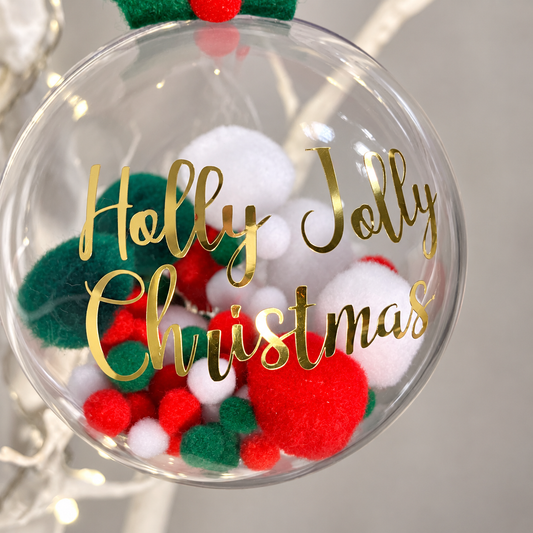 Clear Christmas bauble with red, white, and green pom-poms inside. Gold text reads "Holly Jolly Christmas".
