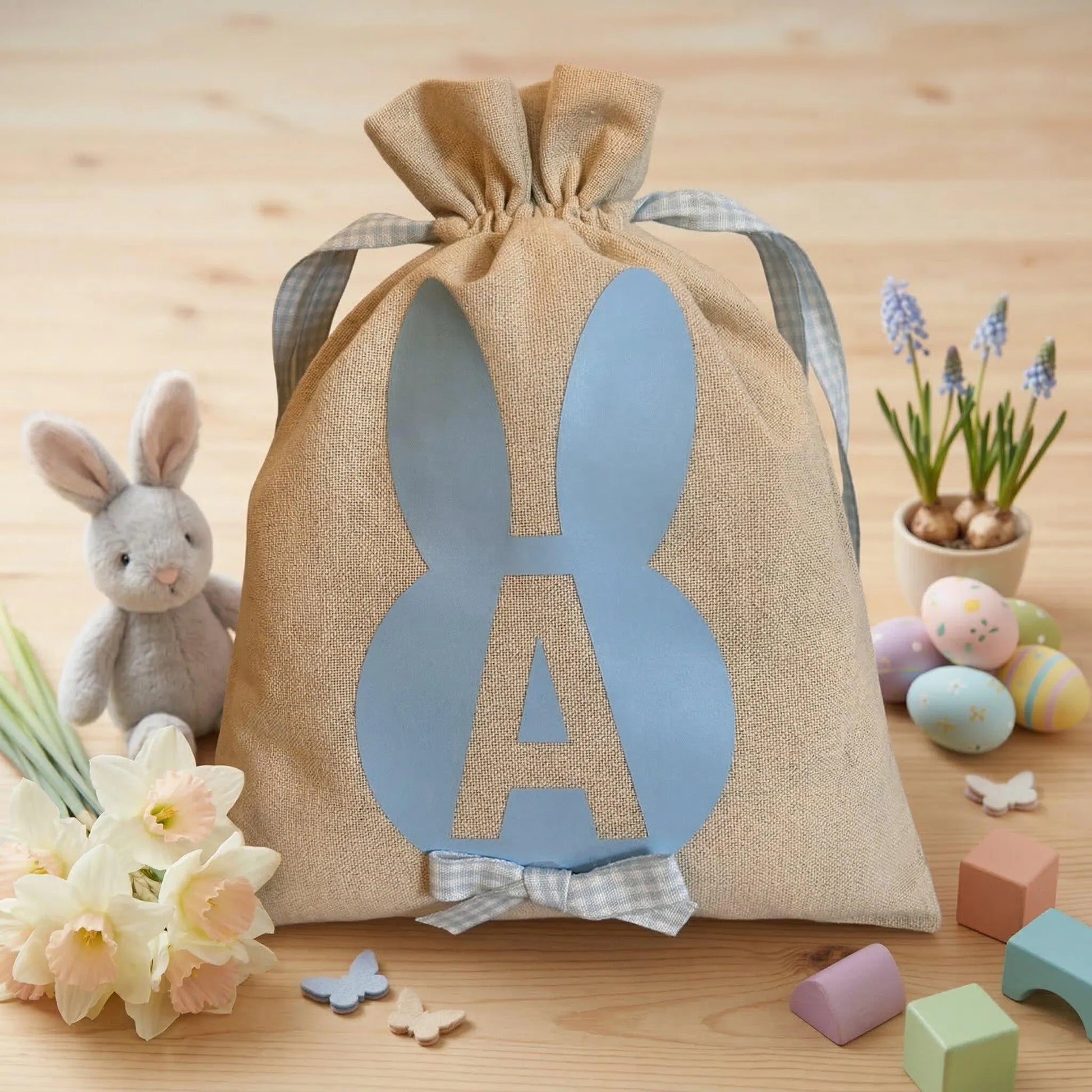 Burlap Easter gift bag with blue bunny ears and letter A, surrounded by a plush bunny, flowers, and eggs