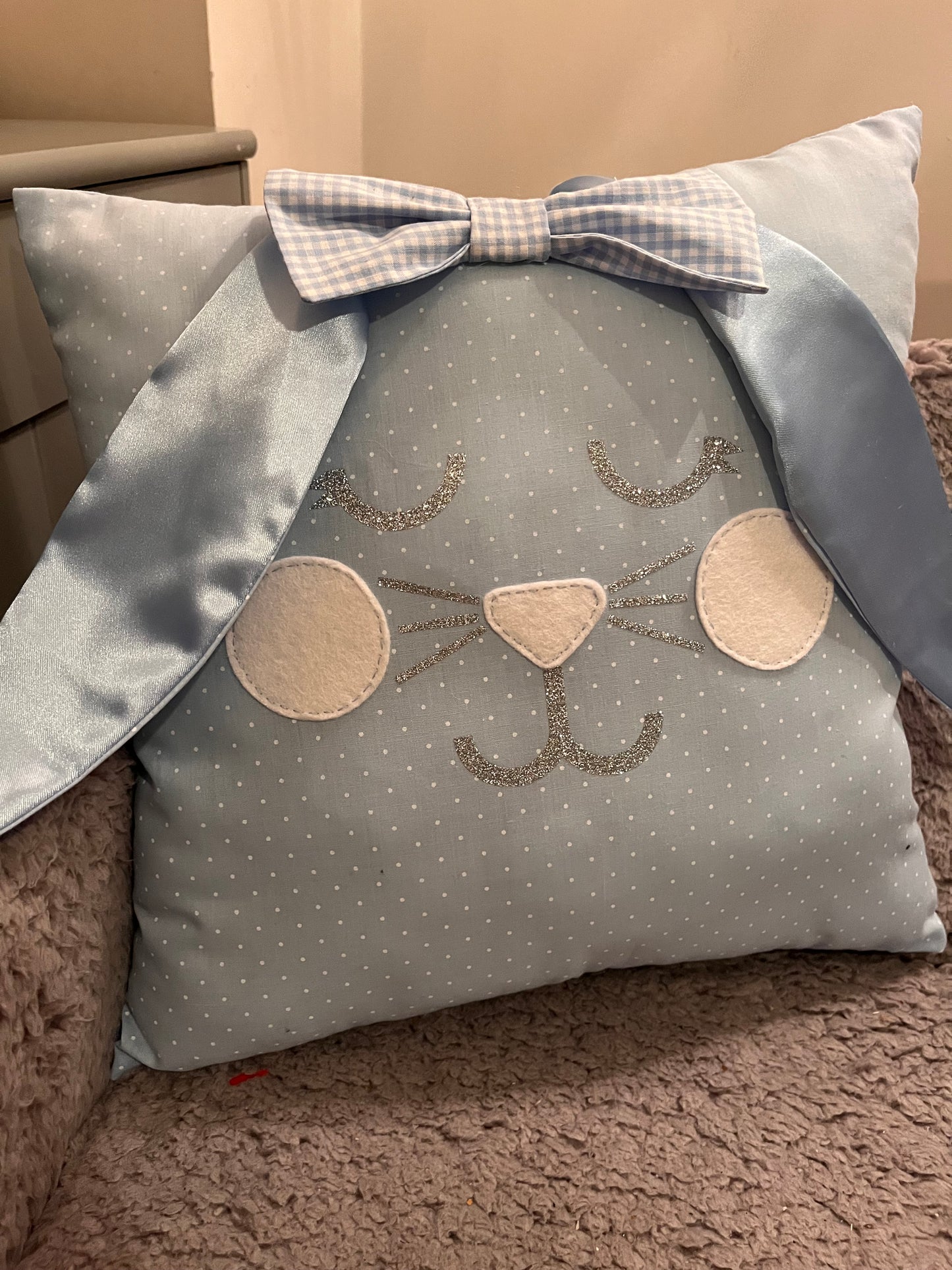 Light blue polka dot pillow with a bunny face, glittery whiskers, and a gingham bow.