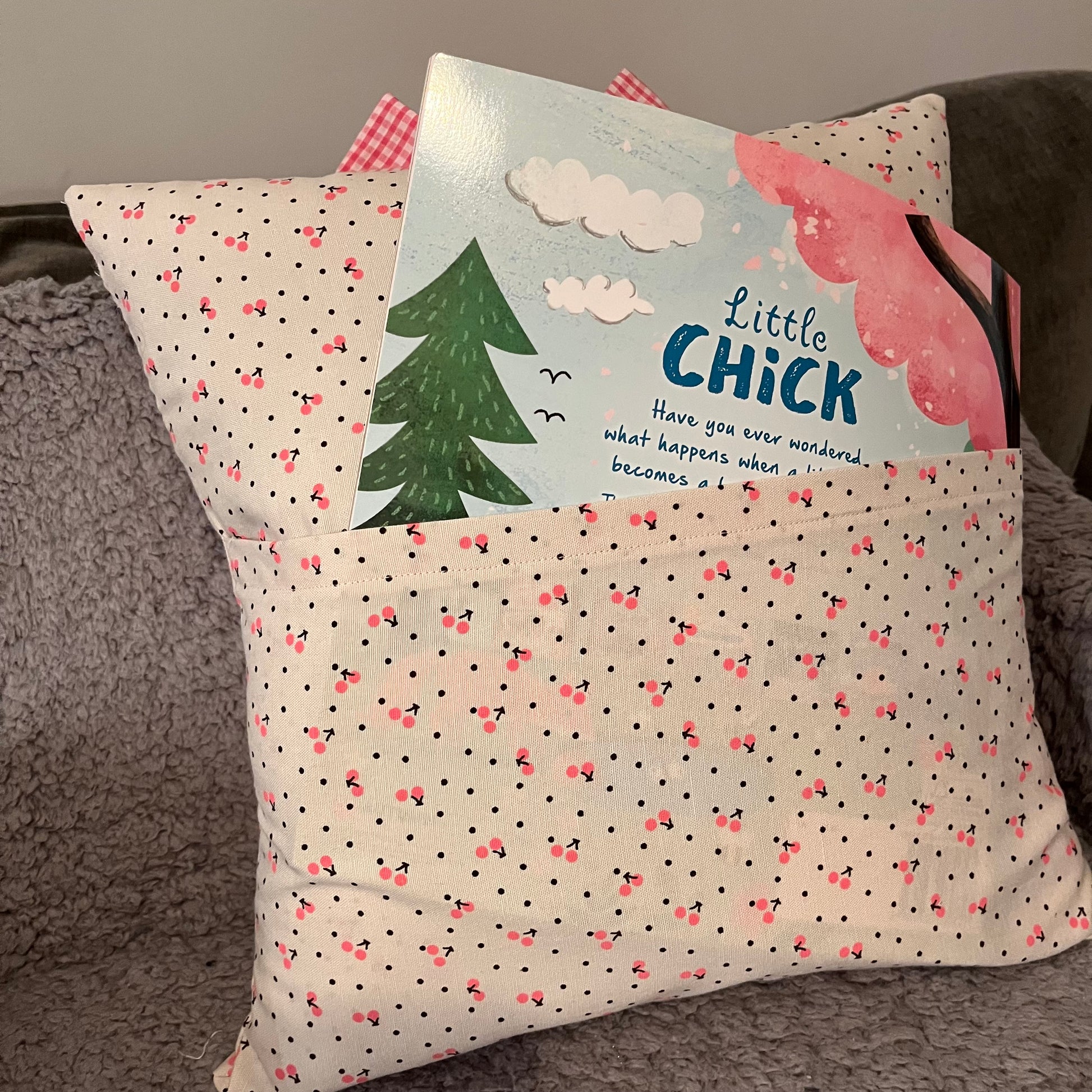 A white pillow with pink cherries and black dots, holding a book titled Little Chick in its front pocket.