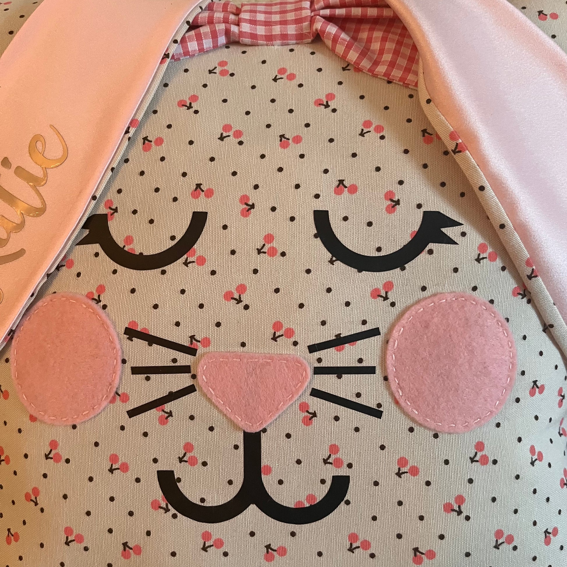 Close-up of a fabric bunny face with pink cheeks, nose, black whiskers, and eyelashes. It has pink cherry and black polka dot patterns. Pink satin ears and a pink gingham bow are visible at the top.